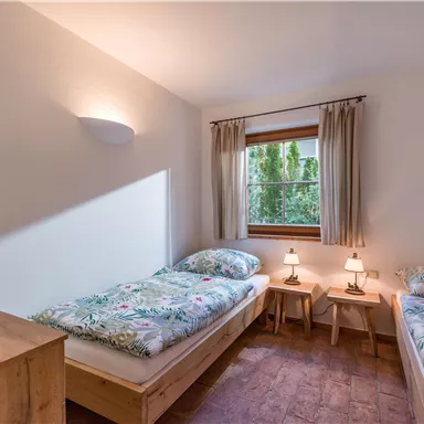 Ein helles Schlafzimmer mit zwei Einzelbetten und gemütlicher Bettwäsche. Am Fenster stehen zwei Nachttische mit Lampen und ein Holzschrank ist im Raum.