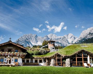 Ein malerisches Landhaus umgeben von Bergen und blauen Himmel. Die blühenden Fensterbänke und grüne Dächer verleihen dem Ort einen charmanten Charakter.
