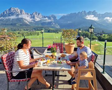 A family is sitting at a table outdoors, enjoying breakfast with a view of the mountains. The landscape is green and sunny, with blooming plants and a clear sky.