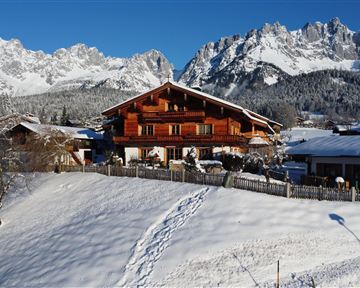 Ein charmantes Holzhaus liegt auf einem schneebedeckten Hügel. Im Hintergrund erstrecken sich majestätische Berge unter einem klaren blauen Himmel.