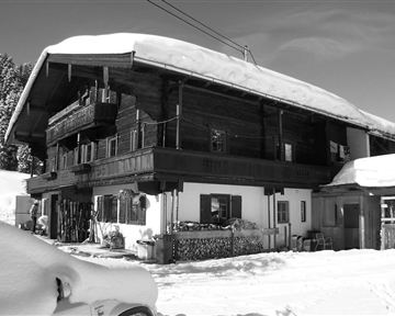 A traditional chalet in the snow with a large, sheltered roof. The wooden facade and balconies give the building a rustic charm.