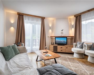 A modern living room with a cozy sofa, a wooden table, and a television. Large windows let in a lot of light and give the room an inviting atmosphere.