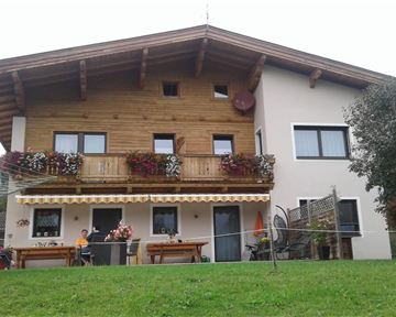 A large house with a wooden balcony and colorful flower boxes. The garden is green and inviting, with some seating options.
