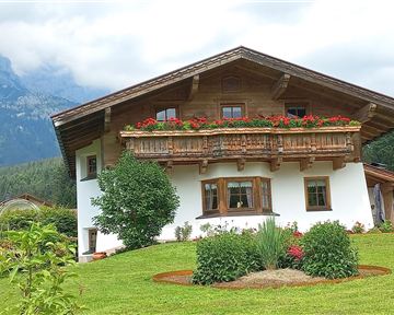 A beautiful, traditional house with a wooden balcony and colorful flowers in the garden. In the background, mountains and a green landscape can be seen.