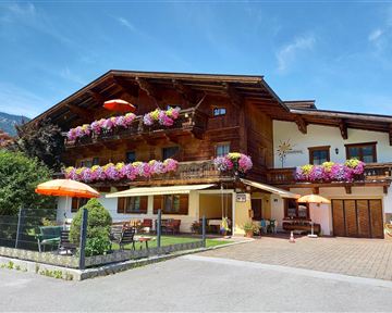 Ein traditionelles Holzhaus mit bunten Blumenbalkonen und einem klaren blauen Himmel. Vor dem Haus befinden sich ein Gartenbereich und Sonnenschirme.