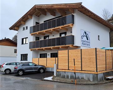 Ein modernes Gebäude mit mehreren Balkonen und einer Holzverkleidung. Vor dem Gebäude stehen einige Autos geparkt.