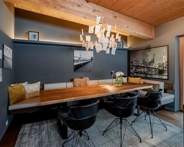 A modern dining room with a large wooden table and black chairs. The walls are painted in a dark color, and a striking chandelier provides lighting.
