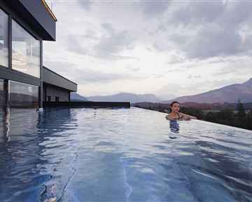 Ein moderner Infinity-Pool mit Blick auf Berge und einen bewölkten Himmel. Eine Person entspannt sich im Wasser.