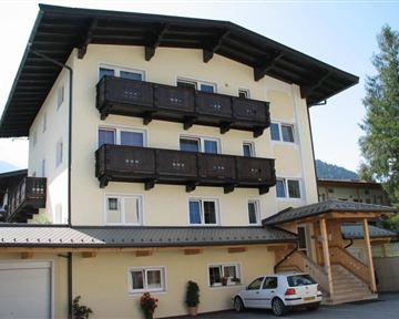 Ein mehrstöckiges Gebäude mit einem hellen Anstrich und Balkonen aus Holz. Vor dem Haus steht ein Auto und es gibt einige Pflanzen im Eingangsbereich.