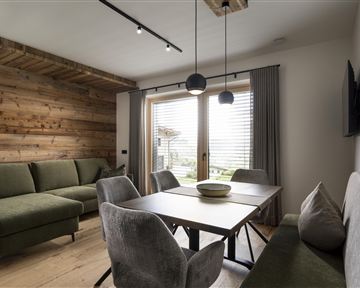 Ein stilvolles Wohnzimmer mit einer grünen Couch und einem modernen Esstisch. Große Fenster lassen viel Licht herein und bieten einen Blick nach draußen.