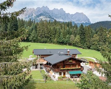 Das Almchalet Weberhof umgeben von dichten Wäldern und Wiesen. Im Hintergrund erheben sich majestätische Berge unter einem klaren Himmel.