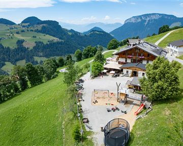 Eine malerisches Hotel umgeben von grünen Wiesen und Bergen. Im Vordergrund ist ein Spielplatz zu sehen.