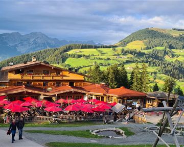 A cozy alpine inn with red sun umbrellas and many guests. In the background, green hills and mountains can be seen.