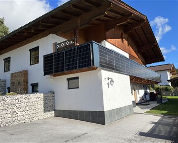 A modern house with a wooden overhang and a balcony. The facade features solar panels and there is a garden view in the background.