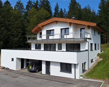 A modern, two-story house in a nature-friendly environment. It has a wooden roof overhang and appears bright and inviting.