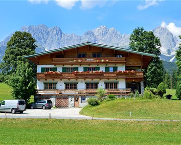 A family-friendly alpine house with flower balconies in a green meadow. In the background, the impressive mountains are visible.