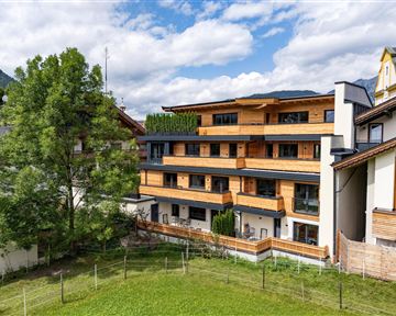 Ein modernes Gebäude mit Holzverkleidung und mehreren Balkonen. Im Vordergrund ist ein grüner Rasen mit einigen Bäumen zu sehen.