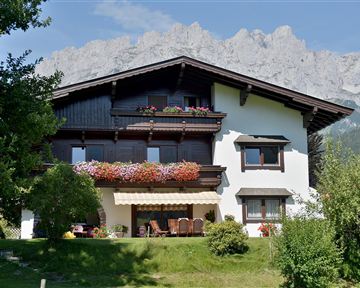 Ein schönes Haus mit blühenden Balkonblumen und einem großen Garten. Im Hintergrund sind beeindruckende Berge zu sehen.