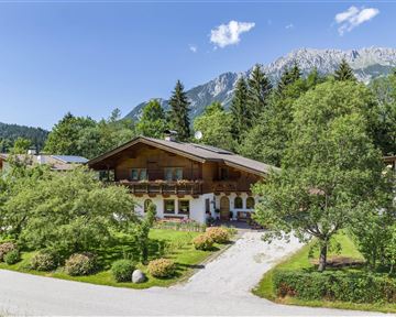 Ein malerisches Haus umgeben von Bäumen und Grünflächen. Im Hintergrund sind majestätische Berge und ein klarer Himmel zu sehen.