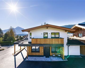 Ein modernes Haus mit Holzverkleidung und großen Fenstern. Im Hintergrund sind Berge und blauer Himmel sichtbar.
