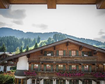 A traditional wooden house with colorful flower balconies and a view of the mountains. The sky is cloudy and the surroundings are green and hilly.