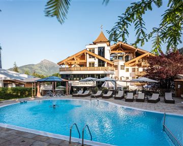 Ein stilvolles Hotel mit einem einladenden Pool und Liegen. Im Hintergrund sind die Berge und ein klarer Himmel zu sehen.
