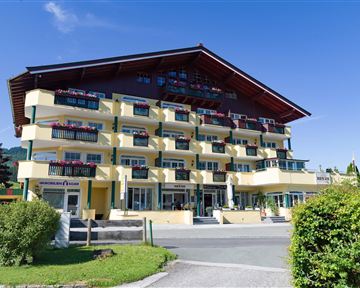 Ein modernes Hotelgebäude mit mehreren Balkonen und bunten Blumen. Es liegt in einer grünen Umgebung unter einem klaren blauen Himmel.