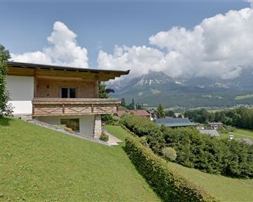 Ein schönes Haus auf einem Hügel mit Blick auf die Berge. Die Landschaft ist grün und bewaldet, unter einem klaren Himmel mit einigen Wolken.