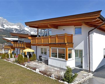 Ein modernes Haus mit Holzverkleidung und großen Fenstern. Im Hintergrund sind Berge und ein blauer Himmel zu sehen.