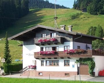 A charming house with a balcony and colorful flowers on the windowsills. In the background, green hills and coniferous trees can be seen.