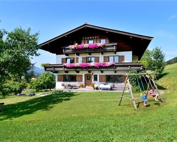 Ein hübsches Bauernhaus mit blühenden Blumenbalkonen und einer großen Wiese. Kinder spielen auf einer Schaukel im Vordergrund.