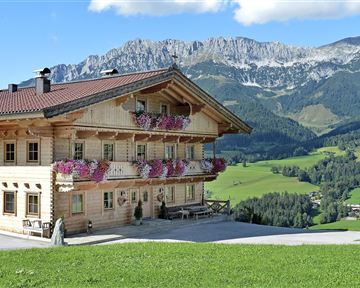 A beautiful wooden house with colorful flower balconies. In the background, green meadows and impressive mountains stretch under a clear sky.