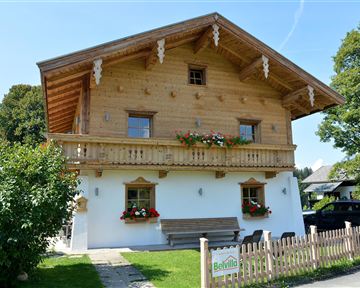 Ein traditionelles, holzverkleidetes Haus mit Balkon und bunten Blumen. Der sonnige Tag verleiht der Szene eine einladende Atmosphäre.