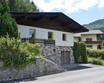 Ein modernes Haus mit einem Steinfußboden und viel Grün. Im Hintergrund sind weitere Gebäude und eine wunderschöne Berglandschaft zu sehen.