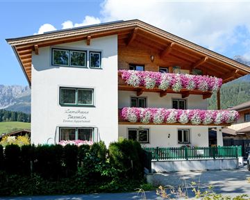 Ein schönes Alpenhaus mit bunten Blumenkästen. Im Hintergrund sind Berge und ein klarer Himmel zu sehen.