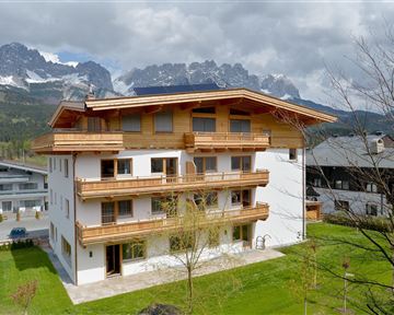 Ein modernes Haus mit Holzverkleidung und Balkonen, umgeben von grüner Wiese. Im Hintergrund sind beeindruckende Berge und ein bewölkter Himmel zu sehen.