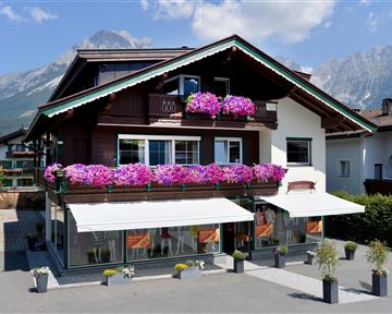 Ein charmantes Holzgebäude mit bunten Blumenkästen und Balkonen. Im Hintergrund sind beeindruckende Berge zu sehen.