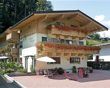Ein gemütliches Holzhaus mit blühenden Balkonen und einer sonnigen Terrasse. Die Umgebung ist grün und einladend.