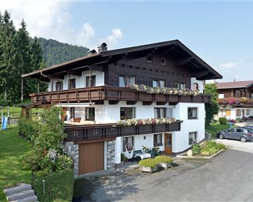 A traditional wooden house in a rural setting with many flowers on the balconies. In the background, green trees and mountains can be seen.
