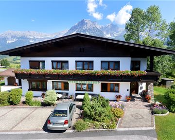 A charming house with flower windows and a carport. In the background, majestic mountains and a blue sky can be seen.