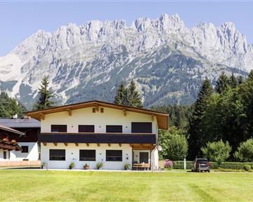 Ein modernes Haus mit einer Wiese und Bäumen im Vordergrund. Im Hintergrund erheben sich beeindruckende Berge unter einem klaren Himmel.