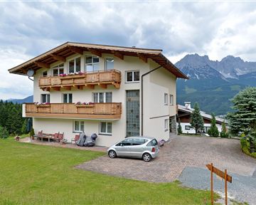 A modern house with balconies and a green lawn area. In the background, mountains are visible under a cloudy sky.