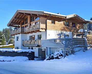 A modern, multi-story wooden house in a snowy landscape. Surrounded by trees and clear blue sky.