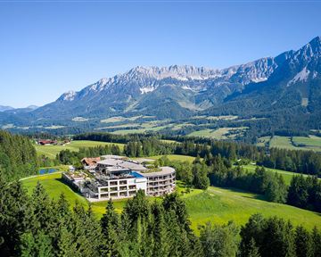Ein modernes Hotel inmitten einer wunderschönen Berglandschaft. Grüne Wiesen und hohe Berge umgeben die Unterkunft.