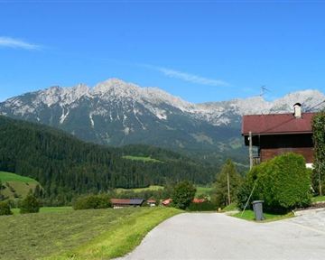 Eine malerische Landschaft mit hohen Bergen und üppigem Grün. Im Vordergrund steht ein schönes Haus an einer ruhigen Straße.