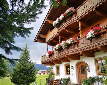 A traditional chalet with blooming balconies and a beautiful garden. Gentle hills can be seen in the background.