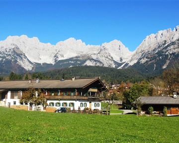 Ein schönes Bergpanorama mit schneebedeckten Gipfeln. Im Vordergrund stehen mehrere Bauernhäuser und eine grüne Wiese.