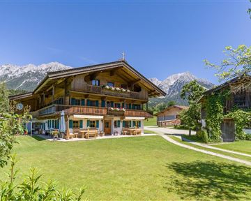 A traditional Alpine house surrounded by green grass and mountains in the background. The sky is clear and sunny.