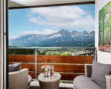 Ein gemütlicher Balkon mit zwei Sesseln und einem Tisch. Im Hintergrund erstrecken sich majestätische Berge unter einem teilweise bewölkten Himmel.