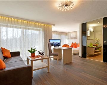 Ein modernes Wohnzimmer mit gemütlichem Sofa, einem Tisch und großen Fenstern. Die orangefarbenen Kissen und Pflanzen verleihen dem Raum eine einladende Atmosphäre.
